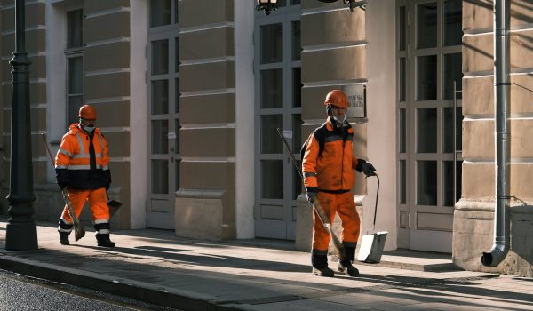 Two Men in Orange Workwear Walking on Sidewalk Holding Broomsticks and Dustpans